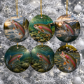 Six round ceramic Christmas ornaments featuring colorful trout fishing scenes, hanging from gold strings against a snowy, frosted tree background.